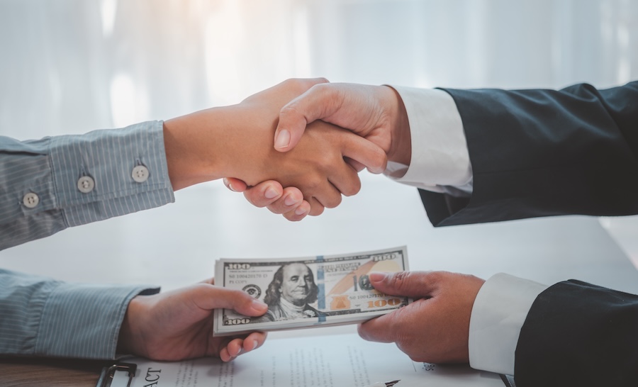 Two people shaking hands over a table, with one holding a stack of hundred-dollar bills, symbolizing a financial or business agreement.