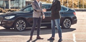 A car sales agent closing a deal with a customer and shaking his hands with a car in the background.