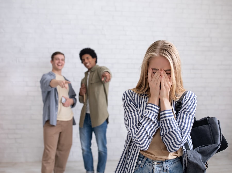 Two individuals pointing and laughing in the background while a young woman in the foreground covers her face, appearing distressed. Workplace bullying concept.