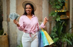 Cheerful woman in casual and hat with many bags of purchases hold dollars.