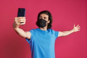 White man in face mask taking selfie on cellphone and showing copyspace.