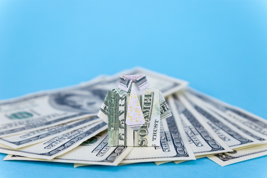 Origami shirt made from a dollar bill placed on a stack of $100 bills against a blue background.