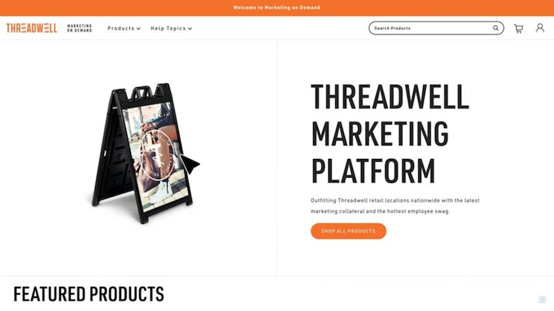 Smartpress Marketing on-demand platform shows a webpage with the company Threadwell as the example. there's a hero image of a sandwich board for the company and to the right of it the headline “Threadwell Marketing Platform” with the subheader “Outfitting Threadwell Retail locations nationwide with the latest marketing collateral, and the hottest employee swag.”