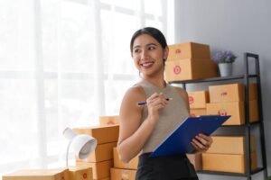Woman holding clipboard smiling near stacked boxes in office. Concept of organization and business planning in a modern workspace.