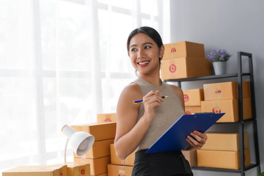Woman holding clipboard smiling near stacked boxes in office. Concept of organization and business planning in a modern workspace.