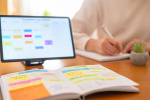 Person writing in a physical daily planner with a digital calendar displayed on a tablet in the background.