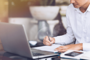 Man in a white shirt working on a laptop and taking notes with a pen in a notebook