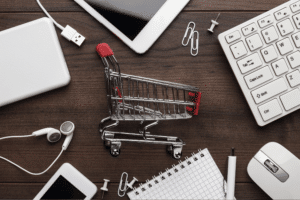 Miniature metal shopping cart surrounded by a white laptop, keyboard, mouse, tablet, and earphones on a wooden desk