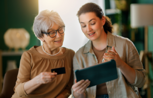A caregiver helping a senior woman use a credit card and tablet to pay for in-home senior care services online.