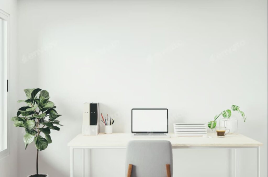 Minimalist home office setup with a white desk, laptop, chair, stacked notebooks, pencil holder, coffee mug, and green plants against a plain white wall.