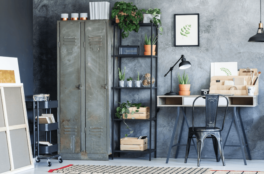 Industrial home office with a wooden desk, black metal chair, vintage metal locker, shelving with plants, desk lamp, and framed botanical art against a textured gray wall, creating a stylish workspace that blends rugged design with natural elements.