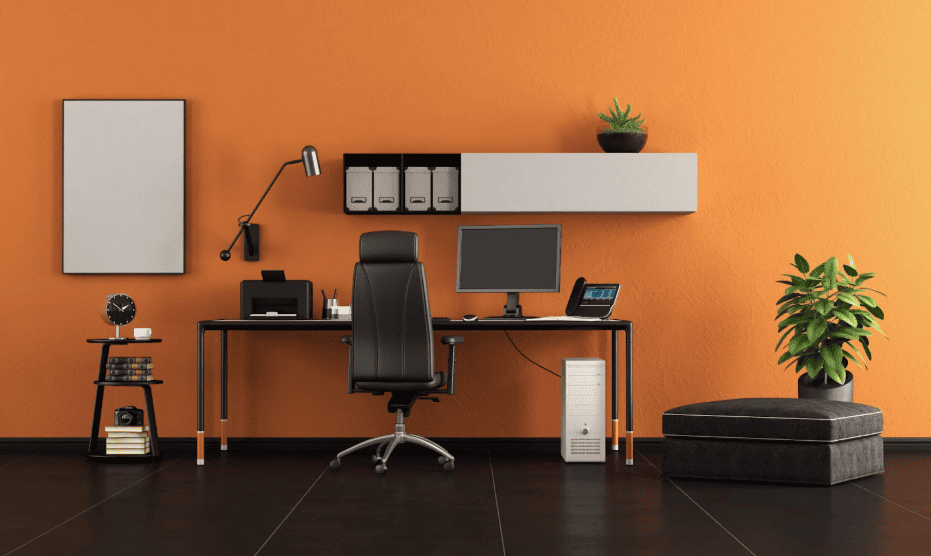 Modern home office with a black desk, computer monitor, ergonomic chair, printer, wall-mounted shelf with storage boxes, desk lamp, and plants against a bold orange wall, creating a clean and tech-friendly workspace.