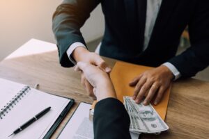 Two corporate businessmen shaking hands while one man places money on document in office room.